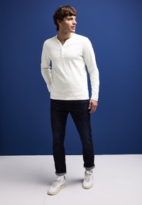 Camisa henley blanca de manga larga con una superficie texturizada, combinada con jeans oscuros y zapatillas blancas. El fondo es azul sólido.