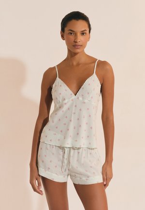 Kobieta ubrana w biały, kwiatowy top na cienkich ramiączkach i dopasowane szorty, stojąca na neutralnym tle.