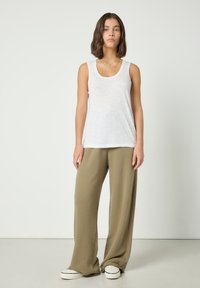 Žena stojí, má na sobě bílý tílko bez rukávů, volné béžové kalhoty a bílé tenisky před jednoduchým světlým pozadím.