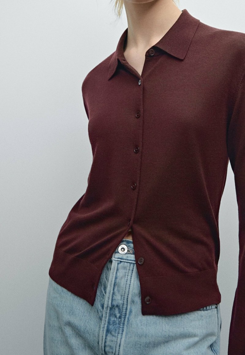 Bordeaux kleurige cardigan met een knoopsluiting aan de voorkant, klassieke kraag en geribde manchetten, gecombineerd met lichtblauwe denim jeans.