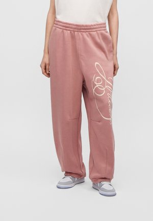 Personne portant un pantalon de jogging rose ample avec un texte blanc script sur la jambe droite et des baskets grises et blanches.