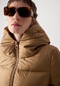 Doudoune marron avec un col montant et une fermeture éclair, présentant une texture lisse et des segments matelassés. Accompagnée de lunettes de soleil surdimensionnées.