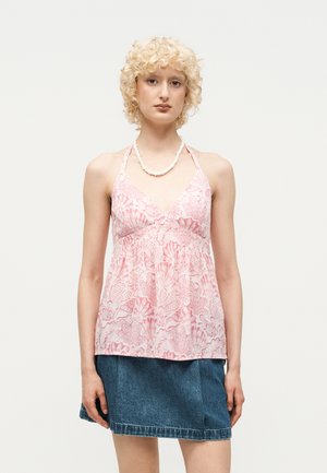 Vrouw met kort krullend blond haar die een roze top met patroon en halternek draagt, een spijkerrok en een witte kralenketting tegen een effen achtergrond.