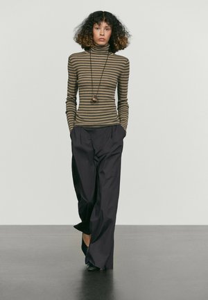 Donna con capelli ricci indossa un dolcevita a righe marroni, pantaloni larghi neri, scarpe nere e una lunga collana con ciondolo, in piedi con le mani nelle tasche.