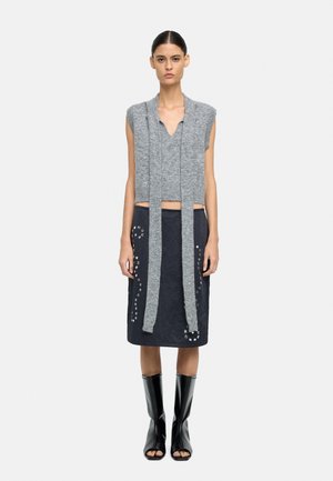 Szary dzianinowy top bez rękawów z długimi, teksturowanymi sznurkami, w połączeniu z ciemną spódnicą z metalowymi przelotkami. Czarne, odkrytoe buty na obcasie.