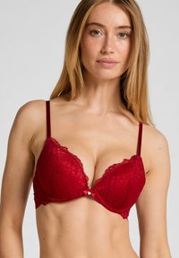 Roter Spitzen-BH mit tiefem Ausschnitt, verstellbaren Trägern und filigranen Blumenmustern. Kleine herzförmige Verzierung in der Mitte vorne.
