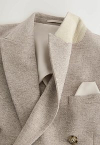 Béžové textúrované sako s výrezom na klopách, svetlo krémovou vnútornou podšívkou, detailom na gombíkoch a zladenou vreckovkou v náprsnom vrecku.