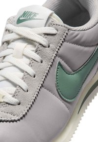 Αθλητικό παπούτσι Nike με πάνω μέρος από γκρι πλέγμα, διακριτικά από σουέντ με υφή, πράσινο δερμάτινο λογότυπο Swoosh, λευκά κορδόνια και πατούσα από καουτσούκ με σχέδιο.