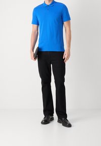 Modré polo tričko s límečkem, krátkými rukávy a třemi knoflíky, v kombinaci s černými kalhotami a černými botami, drželo tablet v levé ruce.