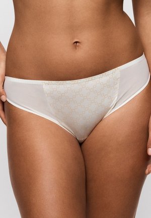 Nainen valkoisessa bikini-tyylisissä alusvaatteissa, joiden etupaneelissa on hienovarainen beige ketjukuosi, yksinkertaista taustaa vasten.