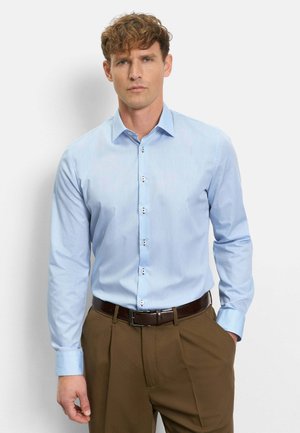 Mann trägt hellblaues, langärmeliges Hemd mit Knöpfen, in braune, plissierte Hosen mit dunklem Gürtel gesteckt, steht vor einfarbigem Hintergrund.