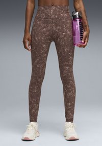 Leggings marron taille haute à motif moucheté, associés à des baskets claires. Le mannequin tient une bouteille d'eau rose transparente.