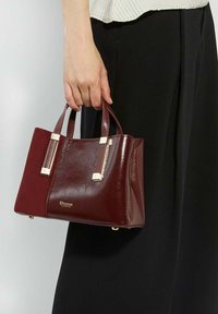Bordeaux leren handtas met glanzend gouden hardware, gestructureerde vorm, dubbele bovenhandvatten en logo-detail aan de voorkant.