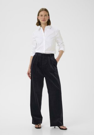 Weißes Hemd mit Knöpfen und hoch taillierte, dunkelblaue Babycord-Hose mit Falten. Das Modell trägt schwarze High Heels. Schlichtes, sauberes Design.