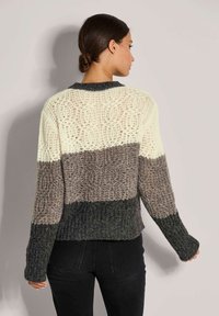 Frau mit dunklen Haaren im Dutt, die einen grob gestrickten, mehrfarbigen Pullover mit cremefarbenen, grauen und dunkelgrünen horizontalen Streifen trägt, steht vor einer schlichten Wand.