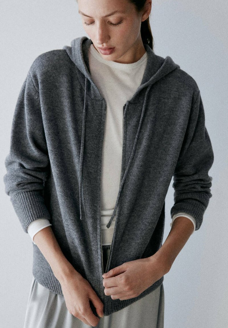 Jeune personne portant un sweat à capuche gris zippé par-dessus un t-shirt blanc à manches longues, regardant vers le bas tout en fermant la fermeture éclair.