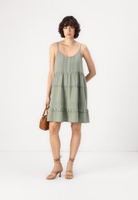 Ανοιχτό πράσινο, αμάνικο φόρεμα με επίπεδη σχεδίαση και φλύνθωτο στρίφωμα, υφασμάτινη υφή και λεπτομερή διακόσμηση στον κορμό. Περιλαμβάνεται καφέ τσάντα.