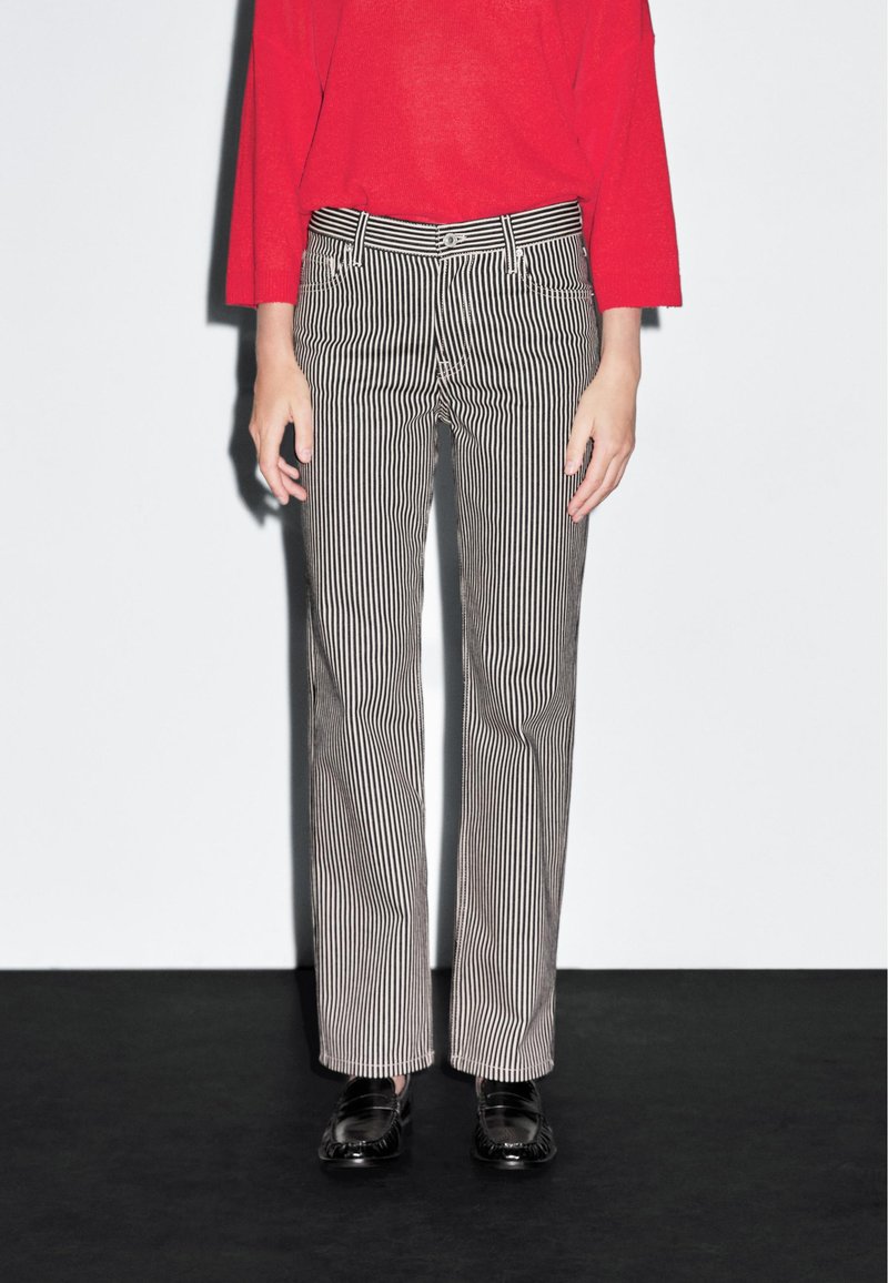 Pantaloni a righe bianche e nere con vestibilità dritta, caratterizzati da un bottone e passanti per la cintura, abbinati a una maglietta rossa a maniche lunghe.