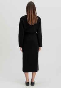 Schwarzes Strickkleid mit langen Ärmeln, tailliertem Schnitt und einem knielangen Saum mit einem seitlichen Schlitz. Glatte Textur mit minimalen Details.