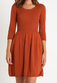 Robe orange rouille avec un corsage côtelé, des manches trois-quarts et une jupe évasée. Texture en maille douce et taille élastique détaillée.