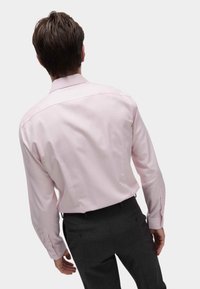 Camicia a maniche lunghe rosa chiaro con colletto e bottoni, in tessuto morbido, abbinata a pantaloni grigi scuri, in una posa in piedi.
