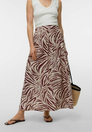 Femme portant un haut blanc sans manches et une longue jupe beige à rayures zèbre marron, tenant un sac en paille et portant des tongs noires.