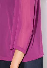 Lila, lös blus med långa ärmar vilande på en arm mot en ljusgrå bakgrund, i kombination med mörka texturerade byxor.