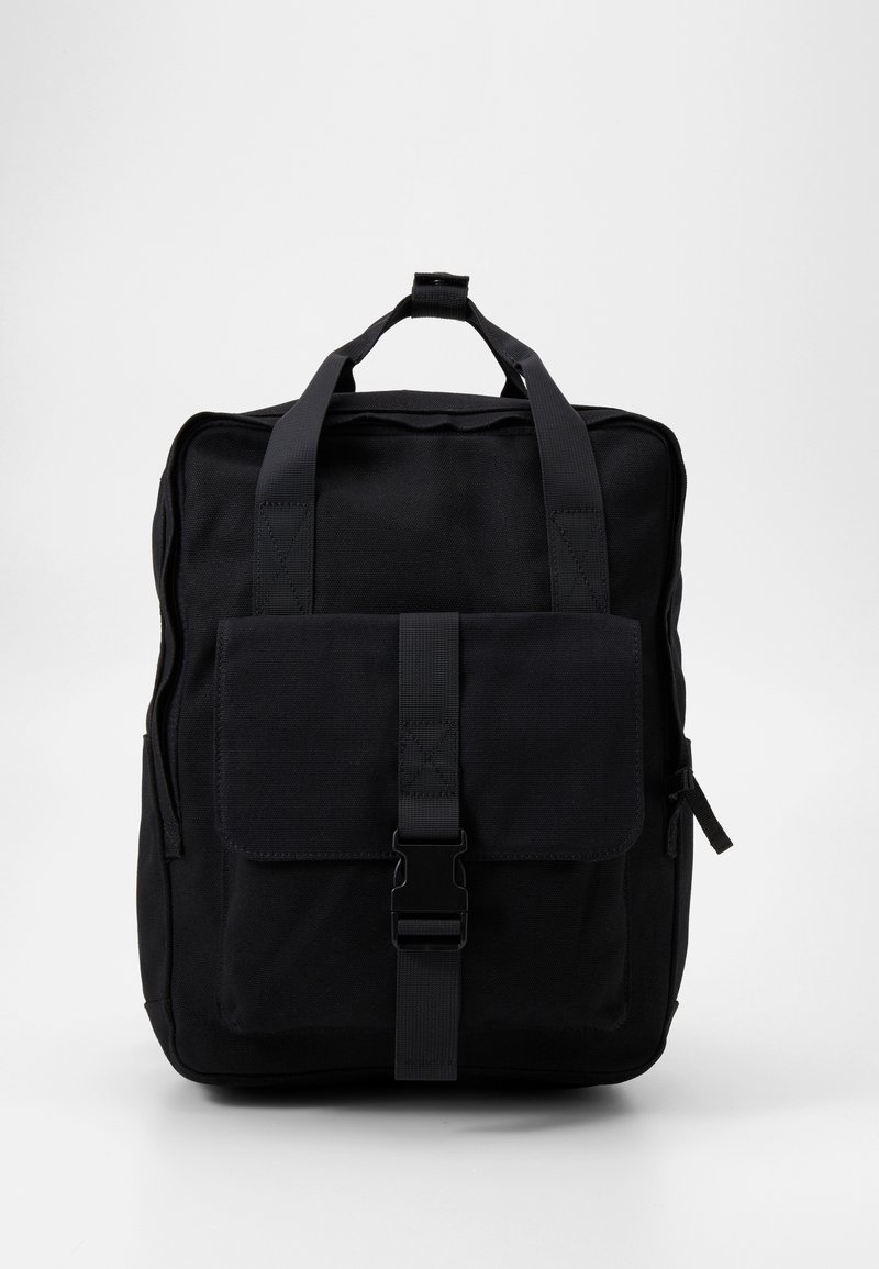 Sac à dos noir en tissu durable, doté de deux poignées supérieures, d'une poche avant avec rabat et fermoir, ainsi que d'une fermeture éclair latérale pour un rangement supplémentaire.