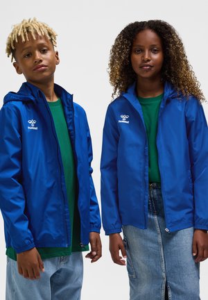 Dwoje dzieci w niebieskich kurtkach Hummel i zielonych koszulkach, stoją obok siebie na jednolitym jasnym tle.