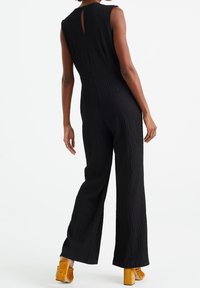 Czarny teksturowany kombinezon z wysokim dekoltem i detalem w postaci dziurki z tyłu. Szerokie nogawki sięgające do kostek. Połączony z żółtymi butami na słupku.