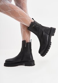 Bocanci negri din piele până la gleznă, cu o talpă groasă, cu închidere prin fermoar pe lateral și textură netedă. Asortați cu ciorapi din plasă albă cu model.