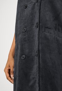 Musta ribbineulosstäinen liivi, jossa pystysuorat uritukset, suuret mustat napit ja sivutasku, korostaen tekstuurista kangasta ja jäsenneltyä muotoilua.