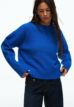 Femme ajustant un pull bleu vif, portant un jean foncé, avec de longs cheveux foncés sur un fond blanc uni.