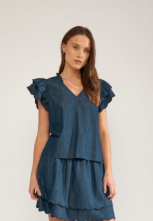 Žena oblečená v tmavomodrý denimový outfit s volánikovými krátkymi rukávmi a viacvrstvovou sukňou s vlnitým lemom, stojaca na jednoduchom pozadí.