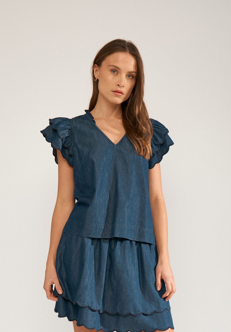 Femme portant une tenue en jean bleu foncé avec des manches courtes à volants et une jupe superposée bordée en feston, debout devant un fond uni.