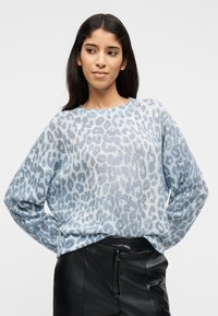 Γυναίκα που φοράει πουλόβερ με ανοιχτό μπλε και λευκό λεοπάρ μοτίβο και μαύρο δερμάτινο παντελόνι, στέκεται με τα χέρια στη μέση και κοιτάζει προς το πλάι.