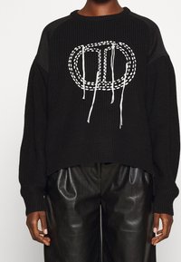 Pull noir en tricot avec une coupe ample et un ourlet côtelé. Il présente un motif brodé blanc et des cordons au niveau de l'encolure.