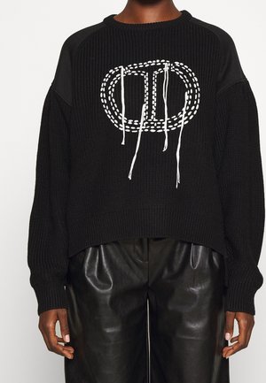 Pull noir en tricot avec une coupe ample et un ourlet côtelé. Il présente un motif brodé blanc et des cordons au niveau de l'encolure.