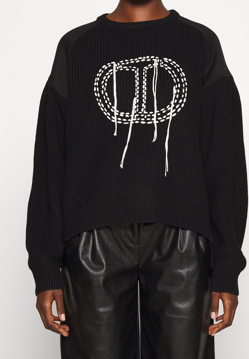 Pull noir en tricot avec une coupe ample et un ourlet côtelé. Il présente un motif brodé blanc et des cordons au niveau de l'encolure.