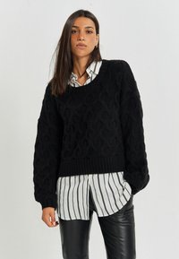 Schwarzer Strickpullover mit strukturiertem Kabelmuster und geripptem Saum, über einem weiß-schwarz gestreiften Hemd mit Knopfleiste getragen.