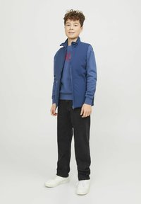 Marinblå zip-up jacka över en blå sweatshirt, kombinerad med svarta jeans och vita sneakers, som visar en avslappnad, lagerpåsatt outfitdesign.