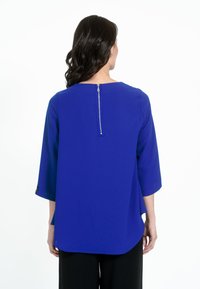 Blusa blu royal con maniche a tre quarti, scollatura rotonda e zip sul retro; il tessuto ha una texture liscia e una vestibilità leggermente loose.