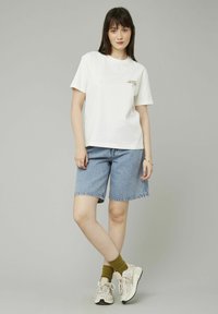 T-shirt en coton blanc à manches courtes, avec du texte imprimé. Porté avec un short en jean bleu et des baskets beiges. Chaussettes vert olive visibles.