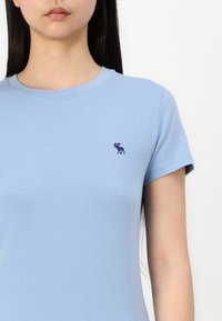 Svijetloplava pamučna majica s okruglim izrezom i kratkim rukavima. Ima mali ljubičasti emblem losa na lijevoj strani prsnog dijela.