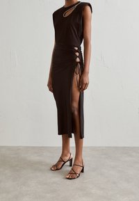 Schwarzes Midikleid mit seitlichen Rüschen, Ausschnitt-Details und hohem Schlitz. Verfügt über kurze Ärmel und eine dezente Akzentuierung am Ausschnitt. Getragen mit Riemchen-Absätzen.