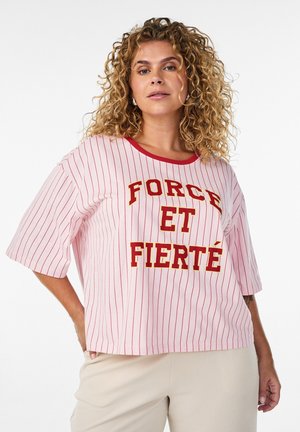 Kvinde med krøllet hår iført en pink og rød stribet trøje med teksten "FORCE ET FIERTÉ", stående med den ene hånd på hoften.