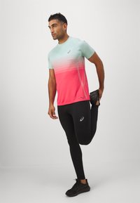 Lett og sporty skjorte med en gradient fra turkis til rosa, med en logo. Sammen med svarte tights og joggesko strekker modellen ut benet.