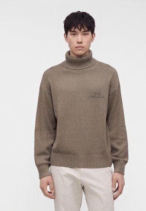 Homme portant un pull à col roulé côtelé marron et un pantalon beige clair, debout devant un fond uni clair.