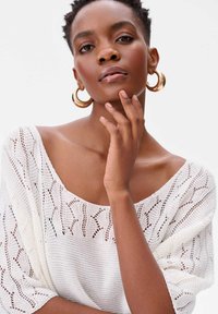 Witte gebreide trui met een open geweven patroon en een brede halslijn, gecombineerd met grote gouden ringoorbellen. Gladde huid en neutrale make-up.