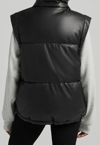 Musta tekonahkapuffervesti, jossa on korkea kaulus, tikattu muotoilu ja hihaton tyyli, kerrostettuna harmaan pitkähihainen paidan päälle.